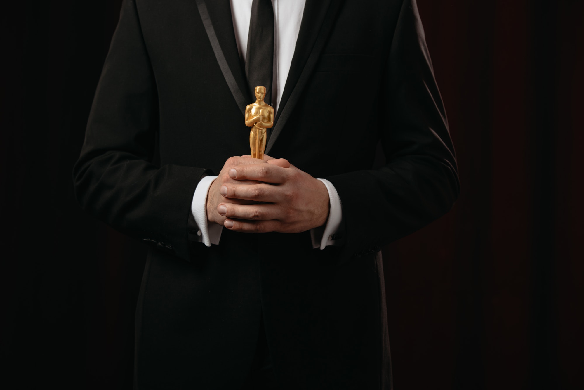cropped view of man in suit holding oscar award on dark background